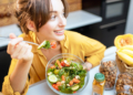 Portrait of a young cheerful woman eating salad at the table full of healthy raw vegetables and fruits on the kitchen at home. Concept of vegetarianism, healthy eating and wellness